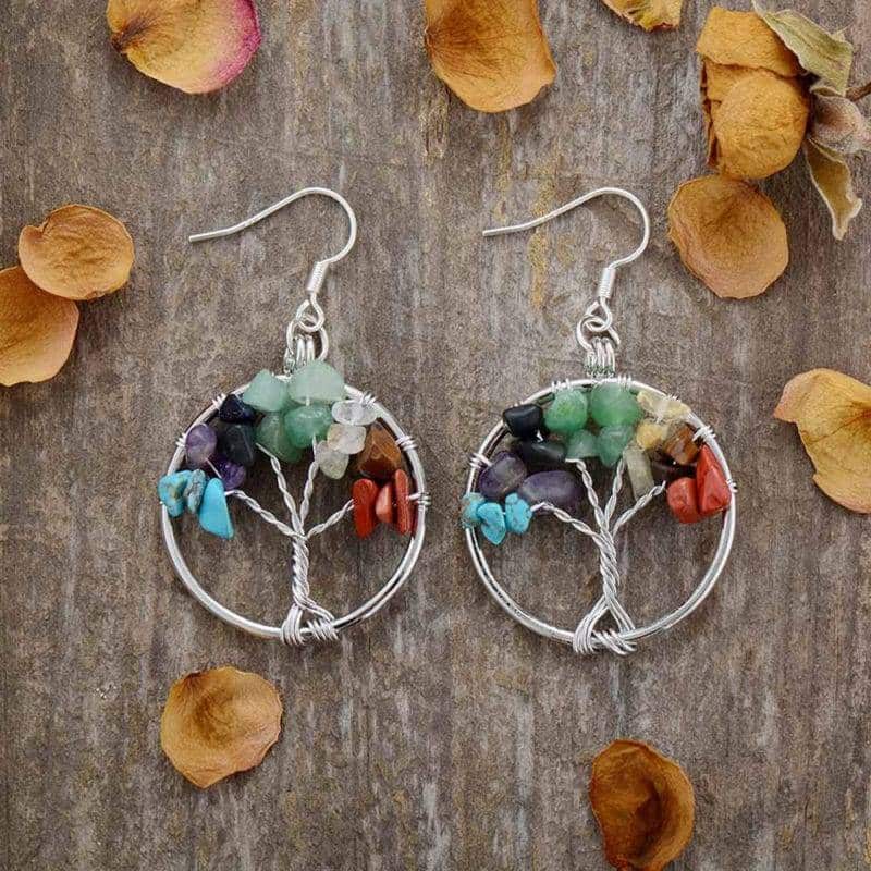 paire de boucles d'oreilles représentant un arbre de vie avec ds pierres de toutes les couleurs entourées par un cercle argent posé sur un fond en bois entouré de pétales