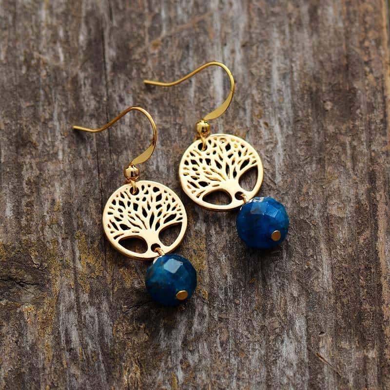 paire de boucles d'oreilles pendantes avec une pierre en forme de perle bleu claire et un arbre de vie argent posée sur un fond en bois