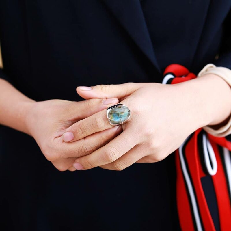 deux mains dont l'une mettant une bague argent avec une pierre bleue sur le doigt de l'autre avec un poignet avec un foulard rouge noir et blanc attaché