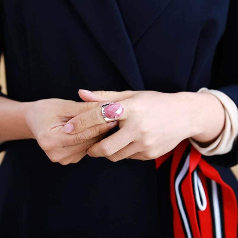 deux mains dont l'une mettant une bague argent avec une pierre rose sur le doigt de l'autre avec un poignet avec un foulard rouge noir et blanc attaché