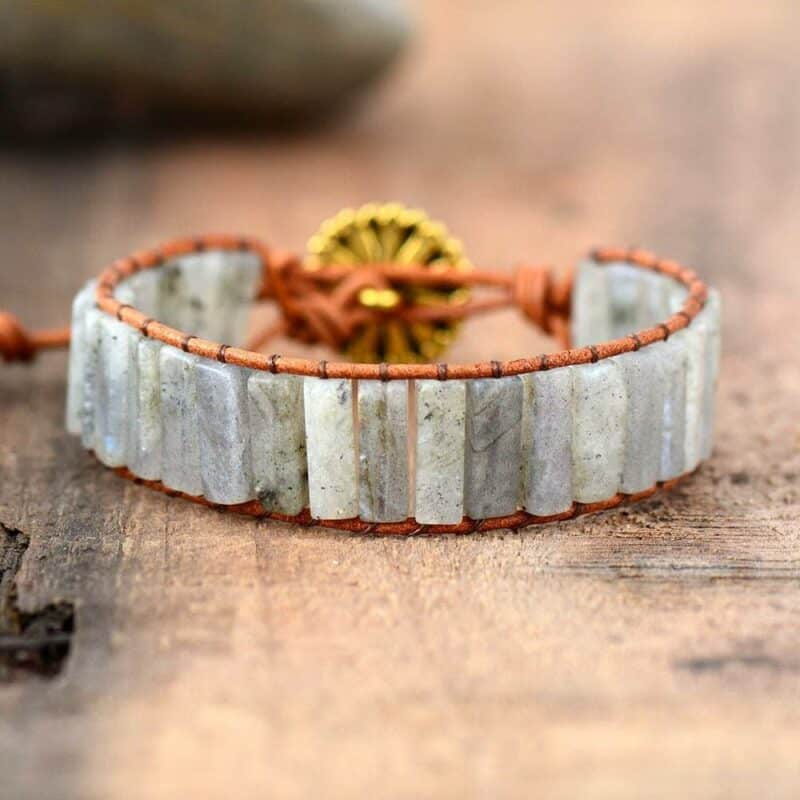 bracelet avec des pierres rectangles de labradorite blanc reliées avec un fil marron et une attache en forme de fleurs posé sur une table en bois