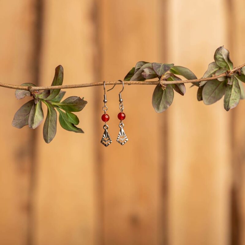 paire de boucle d'oreille pendante avec une pierre en forme de perle rouge et un pendentif argent sur fond bois et pendues à une branche