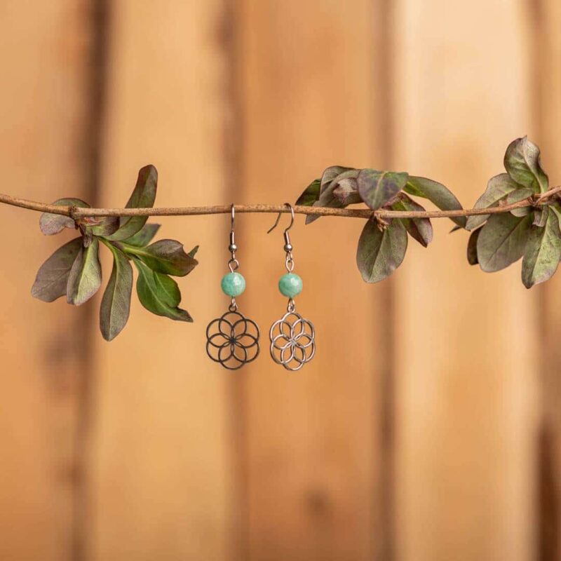 paire de boucle d'oreille pendante avec une pierre en forme de perle bleu et une fleur argent sur fond bois et pendues à une branche