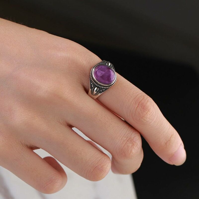 main portant une bague argent avec des gravures et une pierre violette ronde sur fond noir