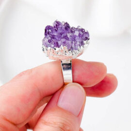 Une bague avec un anneau argenté surplombé d'une améthyste naturelle violette tenu dans une main.