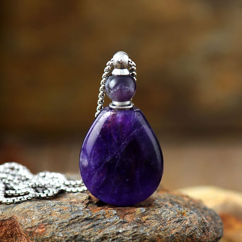 Collier en acier avec pendentif en forme de bouteille de parfum posé sur une pierre sur fond beige flouté