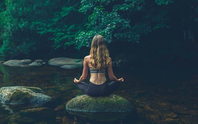 Femme de dos assise en tailleur sur une grosse pierre face à une rivière, elle médite.