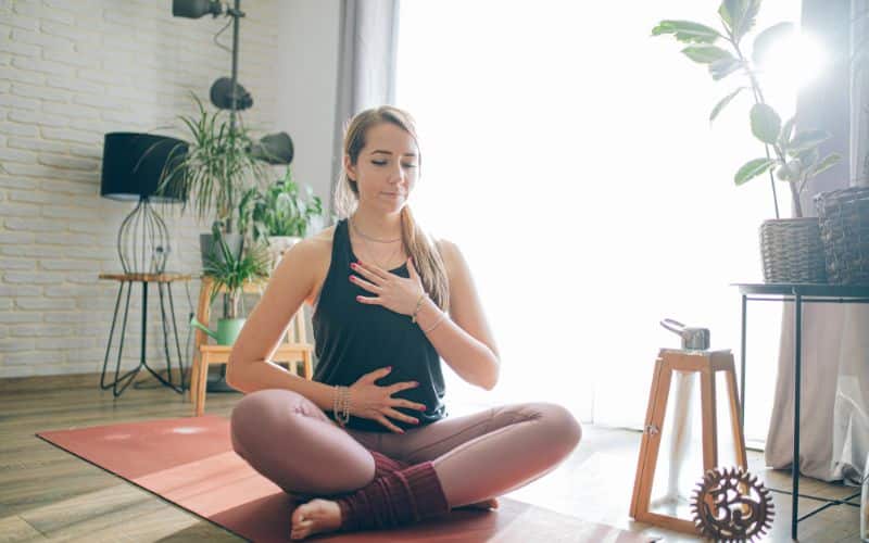 Femme assise en tailleur, sur un tapis de yoga, dans une pièce avec des plantes verte, une main sur le ventre, l'autre sur la poitrine.