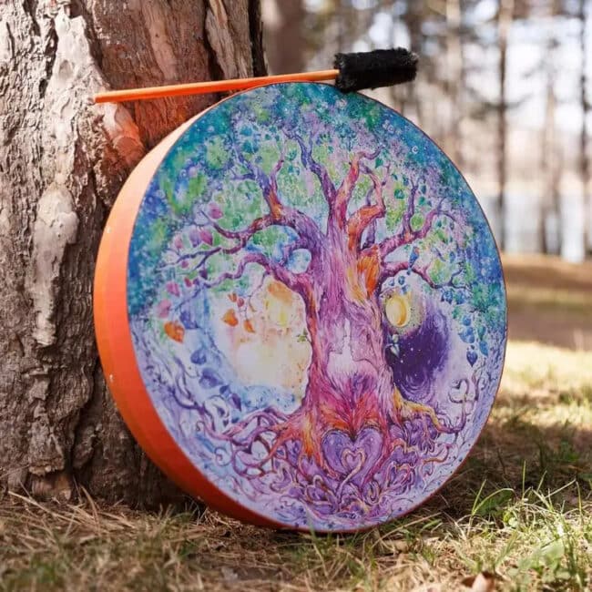 Tambour Chamanique Circulaire au Design d'Arbre d'Amour posé au sol dans l'herbe contre un arbre avec le bâton dessus