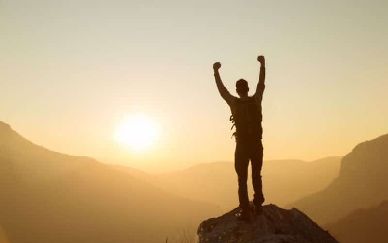 Homme debout sur un rocher face à des montagnes avec le soleil qui se lève. Il a les bras en l'air