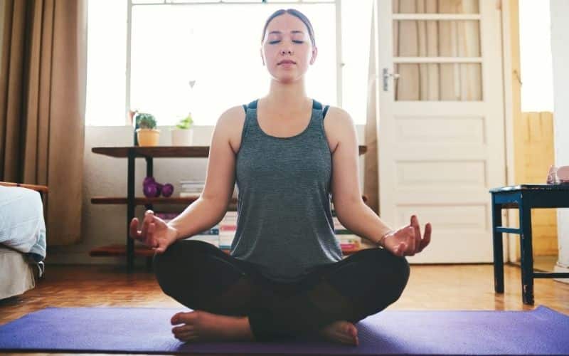 Femme assise en tailleur sur un tapis de yoga, dans une pièce avec une fenêtre derrière elle.