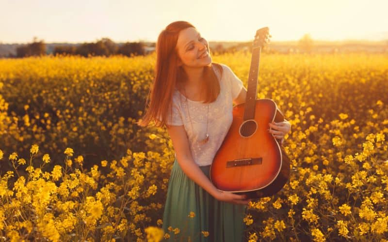 Femme dans un champ de fleurs jaune, portant une guitare avec soleil rasant.