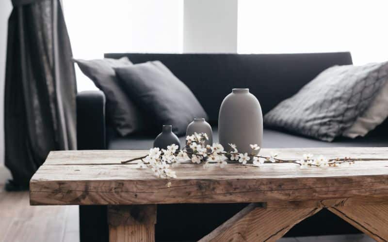 Salon zoomé sur une table basse en bois massif, un canapé et des coussins gris en second plan et une fenêtre et des rideaux en troisième plan.