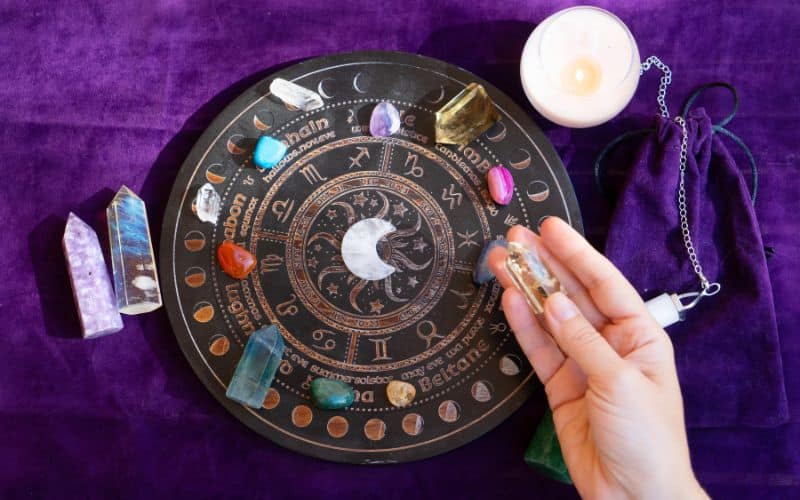 Plateau rond avec les signes astrologiques gravé, les phases de la lune, et les maisons des étoiles, posés dessus des pierres minérales telles que Cristal de Roche, Agathe, Ambre, etc. On voit une main de femme.