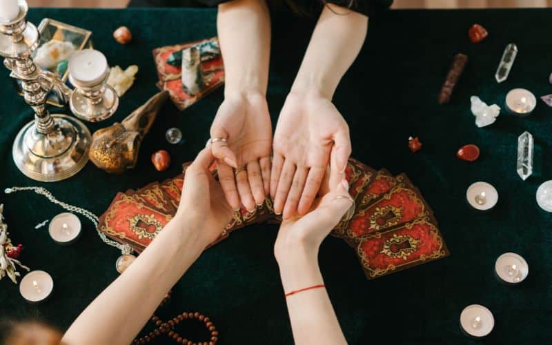 Deux femmes face à face se donnant les mains, paumes vers le haut. Au-dessus d'une nappe noire, avec des bougies, des cartes de tarot, des pierres minérales, etc.
