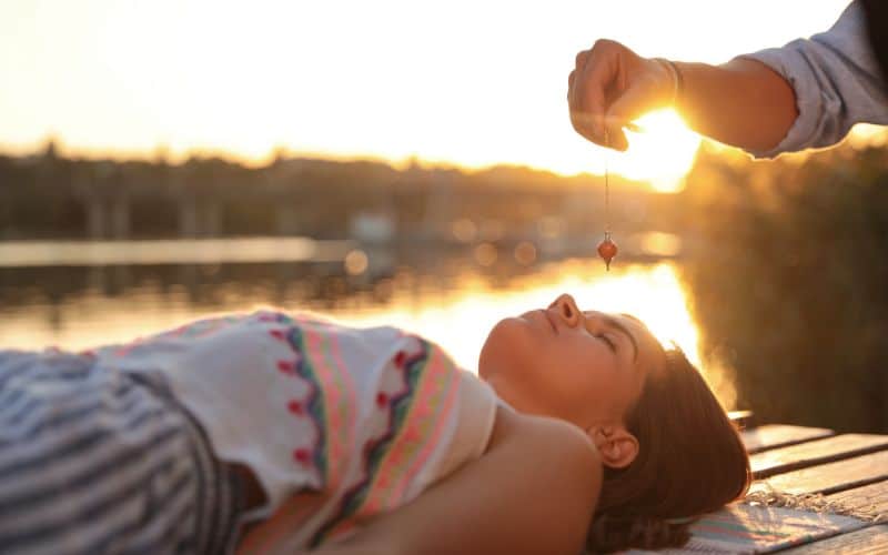 Femme allongée près d'une étendue d'eau avec un soleil couchant. Une personne est assise en tailleur devant elle, et tien un pendule au-dessus de son front.