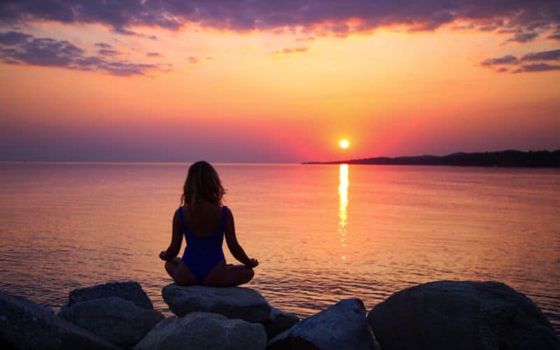 Femme assise en tailleur sur des rochers devant la mer face à un soleil levant.